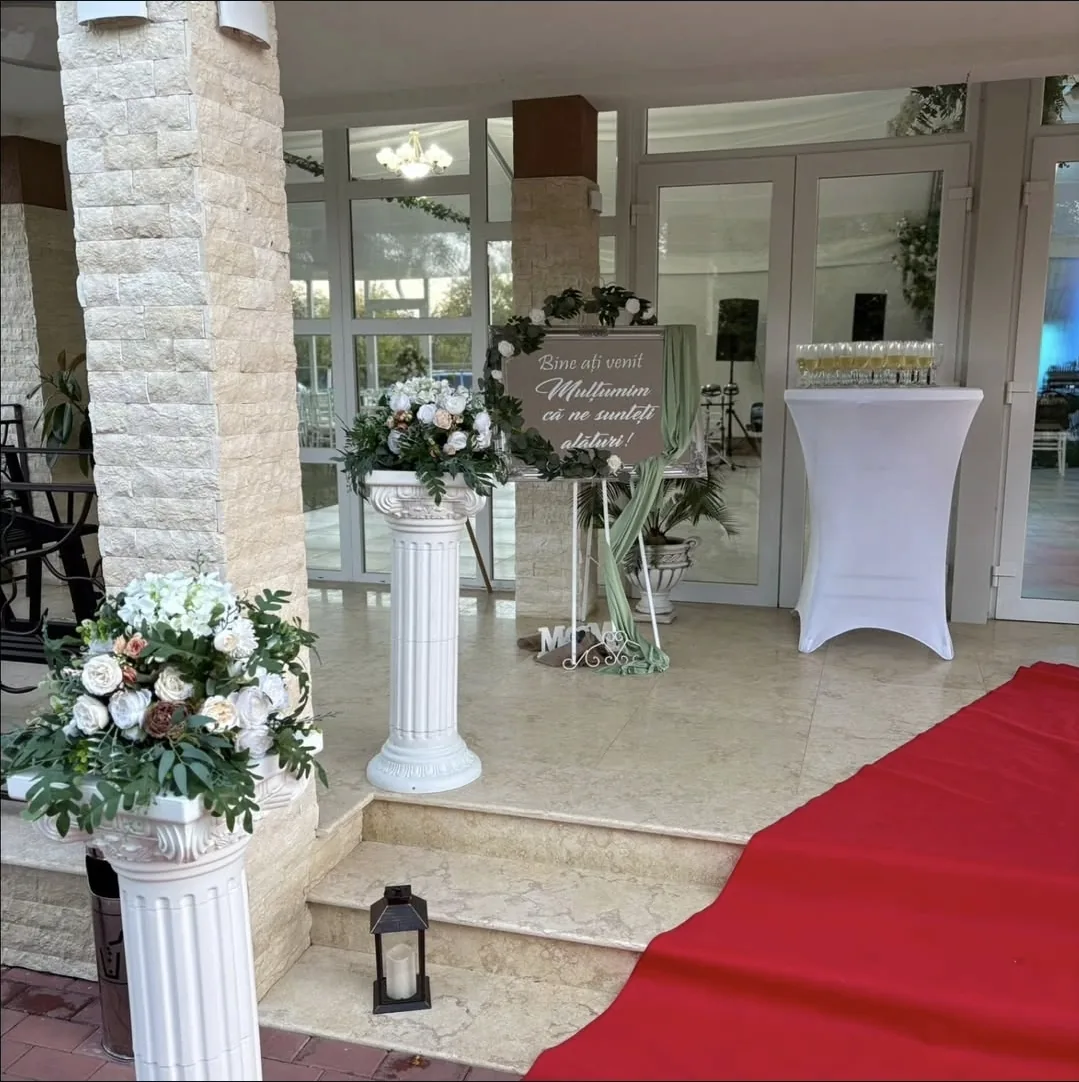 Decor de intrare pentru nuntă cu covor roșu, coloane albe decorate cu aranjamente florale și panou personalizat de bun venit